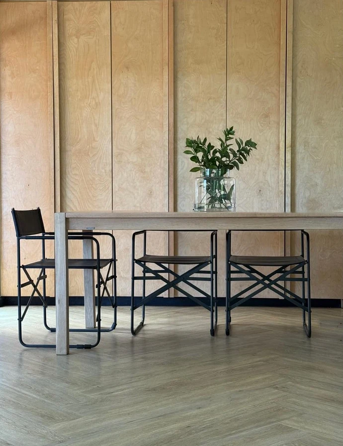 An oak dining table with black folding chairs in front of a wall of birch ply cupboards, a vase of greenery on the table